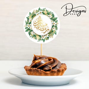 May include: A cupcake with a swirl of caramel frosting sits on a white plate, topped with a round topper. The topper features gold Arabic calligraphy within a white circle, surrounded by a floral wreath of white flowers and green leaves. The text "Designs Drop" is in the upper right corner.