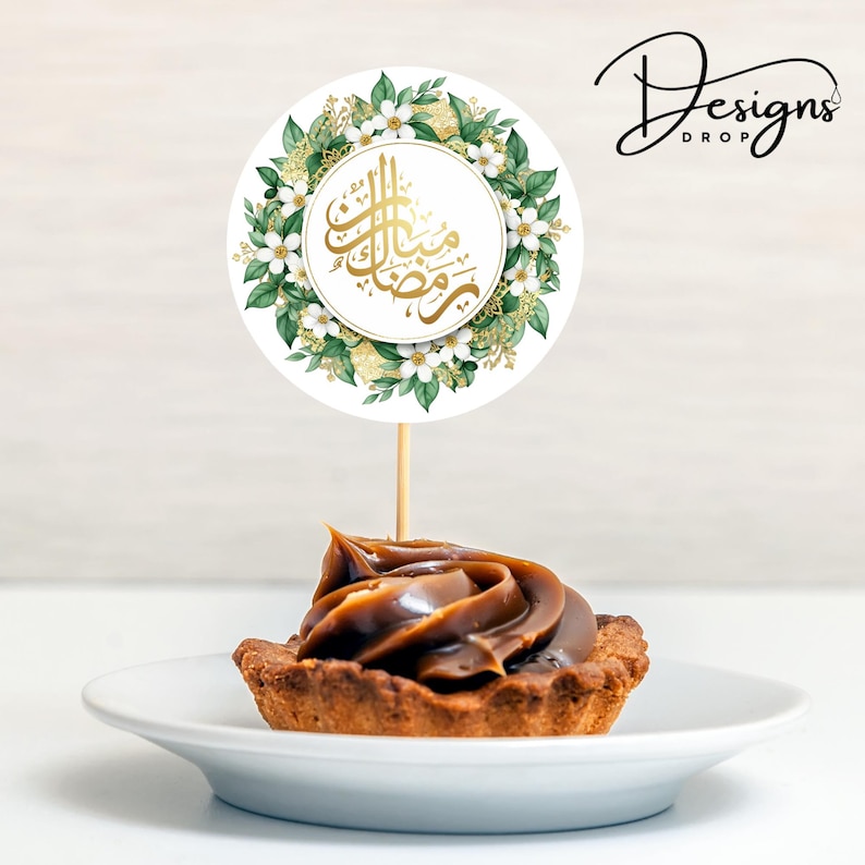 May include: A cupcake with a swirl of caramel frosting sits on a white plate, topped with a round topper. The topper features gold Arabic calligraphy within a white circle, surrounded by a floral wreath of white flowers and green leaves. The text "Designs Drop" is in the upper right corner.
