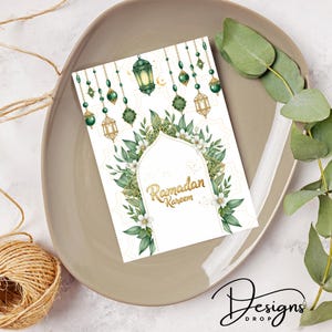 May include: A Ramadan Kareem greeting card with a white background, adorned with green and gold decorative elements, including lanterns and floral designs. The card is placed on a beige plate, with eucalyptus leaves and a twine ball in the background.