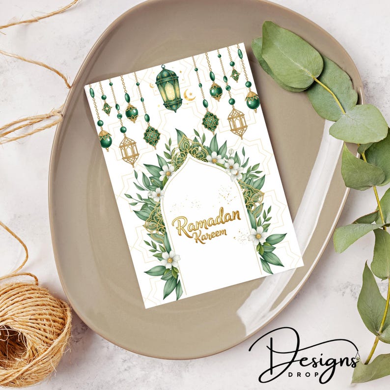May include: A Ramadan Kareem greeting card with a white background, adorned with green and gold decorative elements, including lanterns and floral designs. The card is placed on a beige plate, with eucalyptus leaves and a twine ball in the background.