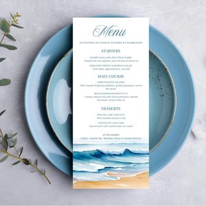 May include: A menu card with a watercolour beach scene at the bottom, placed on a blue plate. The menu lists starters, main courses, and desserts. Gold cutlery and a sprig of greenery are also present.