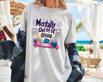 May include: A light gray t-shirt with the words "Mentally Out of Offfice" in a purple and yellow design. The graphic includes a desk with office supplies and a coffee mug. The shirt is worn with a blue and black plaid shirt.