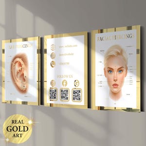 May include: Three framed art prints with gold frames. The prints feature illustrations of ear and facial piercings, and a business card design with contact information and social media icons. The bottom left corner has a gold circle with the text "REAL GOLD ART".