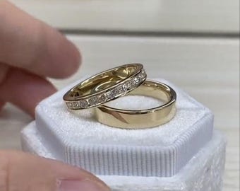 Könnte beinhalten: Zwei goldene Eheringe, einer mit einer Reihe kleiner Diamanten, in einer weißen Ringbox.