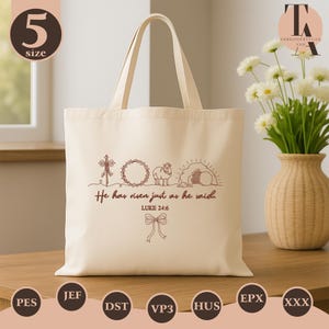 May include: Cream-colored tote bag with a brown embroidered design featuring a cross, wreath, lamb, and tomb. The text "He has risen just as he said. Luke 24:6" is also embroidered on the bag. The bag has two handles.