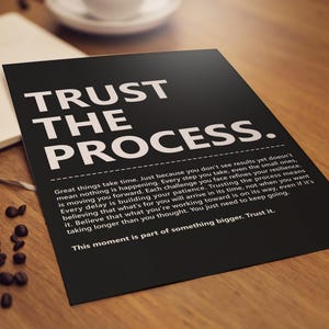 Pode incluir: Um cartaz preto com as palavras "TRUST THE PROCESS" em letras brancas grandes. Abaixo, um texto branco menor fornece uma mensagem inspiradora. O cartaz está sobre uma superfície de madeira, ao lado de um caderno e grãos de café.