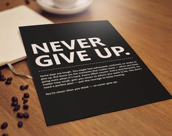 May include: A black poster with the words "NEVER GIVE UP" in large white letters. Below, smaller white text provides motivational advice. The poster is on a wooden surface, with a notebook, coffee beans, and a cup in the background.