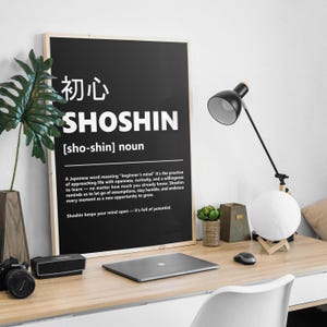 May include: A framed black and white poster with the Japanese word "Shoshin" and its definition. The poster is on a wooden desk with a laptop, lamp, and decorative items. The text on the poster reads: "A Japanese word meaning 'beginner's mind'"