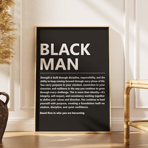 以下が含まれることがあります： 「BLACK MAN」の文字が大きく白抜きで書かれた、額入りの白黒アートプリント。強さ、規律、自尊心についてのモチベーションメッセージが印刷されています。アートワークは木の床に展示されています。