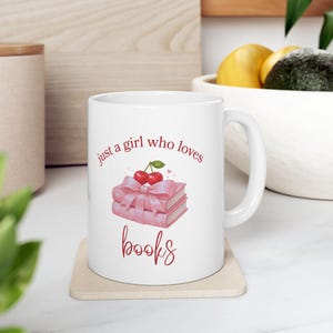 May include: White ceramic mug with the text "just a girl who loves books" in red script. The mug features a watercolor illustration of two pink books tied with a pink bow and topped with cherries. The mug sits on a beige coaster.