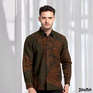 Peut inclure: Une chemise à manches longues vert olive avec un motif batik marron. Le motif batik présente des motifs en spirale et floraux. La chemise est portée par une personne. La marque "JitoArt" est visible en bas à droite. Fond blanc.