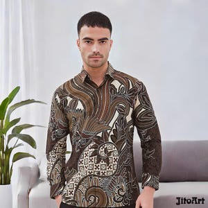 Op de afbeelding: Een shirt met lange mouwen met een bruin en crèmekleurig batikpatroon. Het shirt heeft een gedetailleerd bloemen- en geometrisch ontwerp. Het shirt wordt gedragen door een persoon in een studio.