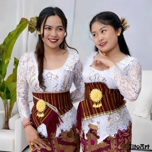 May include: Two women wearing traditional Balinese attire. They are dressed in white lace tops, maroon and gold patterned skirts, and matching maroon and gold belts with gold embellishments. They are smiling and posing for the camera.