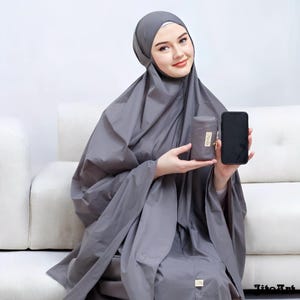 May include: A gray prayer garment set, including a head covering and a matching carrying case. The set is held next to a black smartphone. The carrying case has a gold zipper and a small tag with text.