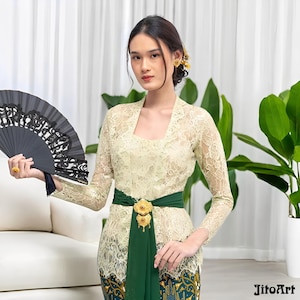 Puede incluir: Una mujer con un top de encaje color crema con mangas largas, una faja verde y una falda estampada. Sostiene un abanico negro con un diseño decorativo. También se ven accesorios dorados para el cabello y un anillo.