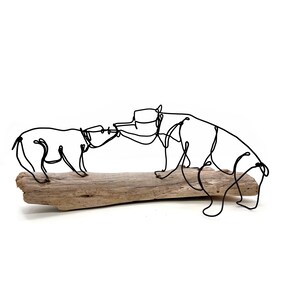 May include: Black wire sculpture of two bears interacting, mounted on a piece of driftwood. The bears are depicted in a continuous line, with one bear standing and the other in a walking position. The sculpture is set against a white background.