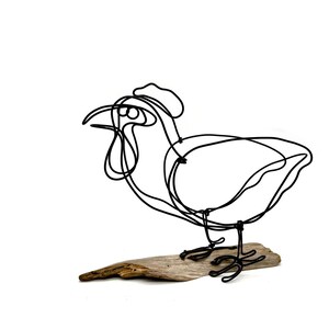 May include: A wire sculpture of a rooster standing on a piece of driftwood. The rooster is made of black wire and is in a simplified, stylized form.