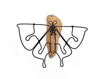 May include: A decorative wall hanging featuring a black metal wire butterfly design mounted on a piece of driftwood. The butterfly's wings have a unique, abstract shape. The driftwood is a light brown color.