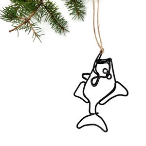 May include: Black wire fish ornament suspended by twine. The fish is a minimalist design with a simple outline. A green pine branch is in the upper left corner, set against a white background.