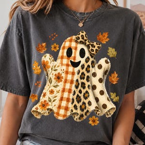 Pode incluir: Uma t-shirt de manga curta cinzento-carvão com um gráfico central de um fantasma com um laço. O fantasma tem secções com xadrez laranja, padrões florais, estampado de leopardo e bolinhas, rodeado por folhas de outono laranja e amarelas.