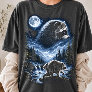 May include: A charcoal gray t-shirt featuring a graphic print of a raccoon in a forest landscape at night. The design includes a large moon, a starry sky, a waterfall, and a second raccoon walking near the water, all rendered in shades of blue and white.