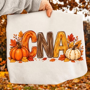 Pode incluir: Uma camisola branca com as letras CNA numa fonte texturizada que imita madeira. O design é rodeado por abóboras cor de laranja e brancas e várias folhas com cores de outono, em tons de vermelho, laranja e amarelo.