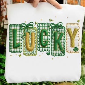 Pode incluir: Saco tote branco com a palavra "LUCKY" bordada em verde e dourado. As letras estão em molduras individuais com xadrez verde e costuras decorativas. Pequenos trevos e estrelas estão espalhados ao redor das letras.