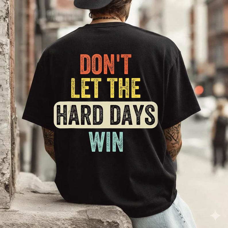 May include: Black t-shirt with the text "DON'T LET THE HARD DAYS WIN" in orange, yellow, and blue. The text is centered on the back of the shirt, with a white rectangular background for "HARD DAYS".