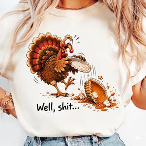 Pode incluir: Uma t-shirt de cor creme com um peru de desenho animado de olhos arregalados a olhar para uma tarte de abóbora caída no chão. O peru tem penas castanhas e cor de laranja. Abaixo do gráfico, o texto diz "Well, shit..." em preto.