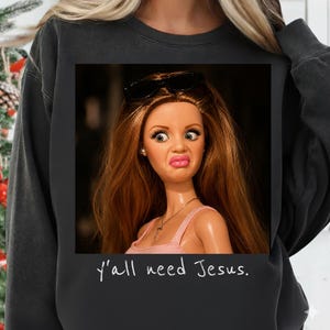 May include: A charcoal gray crewneck sweatshirt featuring a graphic print of a doll with long brown hair, pink lips, and black sunglasses perched on her head. The doll wears a thin gold chain with a cross pendant. The text y'all need Jesus is printed below.