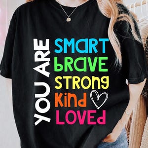 May include: Black t-shirt with the words "YOU ARE" in white, stacked vertically on the left. Below, the words "SMART, BRAVE, STRONG, KIND, LOVED" are in bright colors. A white heart graphic is next to "Kind".
