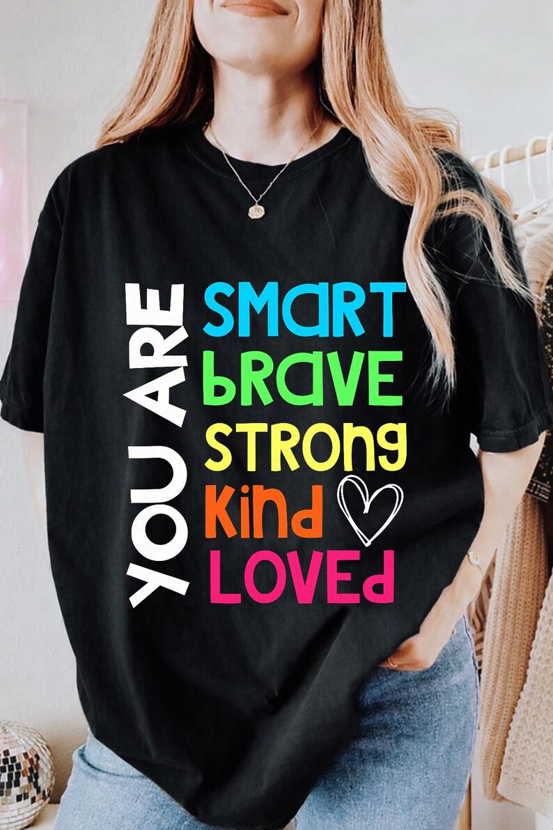 May include: Black t-shirt with the words "YOU ARE" in white, stacked vertically on the left. Below, the words "SMART, BRAVE, STRONG, KIND, LOVED" are in bright colors. A white heart graphic is next to "Kind".