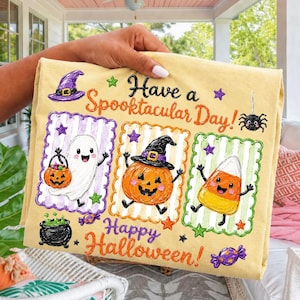Pode incluir: Uma t-shirt amarelo-claro com o texto Have a Spooktacular Day! e Happy Halloween! com ilustrações de um fantasma com abóbora, uma abóbora com chapéu de bruxa, um doce de milho e um caldeirão preto.