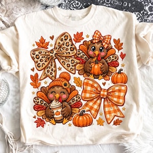 May include: A cream-colored t-shirt featuring a graphic print of two cartoon turkeys, one wearing an orange beanie and holding a coffee cup, the other wearing a plaid bow. The design includes leopard print bows, orange pumpkins, and autumn leaves.