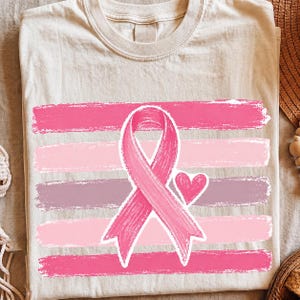 Może przedstawiać: Kremowy t-shirt z grafiką przedstawiającą różową wstążkę świadomości i małe różowe serce, umieszczone centralnie na pięciu poziomych, teksturowanych pasach przypominających pociągnięcia pędzla w odcieniach różu, fioletu i jasnego różu.