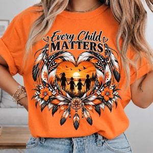 Puede incluir: Una camiseta naranja con un estampado gráfico que incluye el texto Every Child Matters. El diseño presenta una forma de corazón hecha de plumas, flores naranjas y un atrapasueños. Dentro del corazón hay una escena de atardecer con cuatro siluetas de niños.
