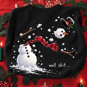 May include: A black sweatshirt featuring a graphic of a snowman falling apart with its head, top hat, and stick arms floating in the air. A red and black plaid scarf is draped around the snowman's neck. The text "well shit..." is printed in white.