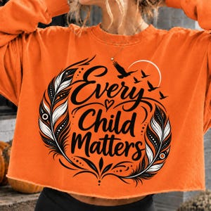 May include: An orange long-sleeve sweatshirt featuring a black graphic print. The design includes two large feathers framing the text Every Child Matters, a small heart, a crescent moon, and several birds in flight, all rendered in black ink.
