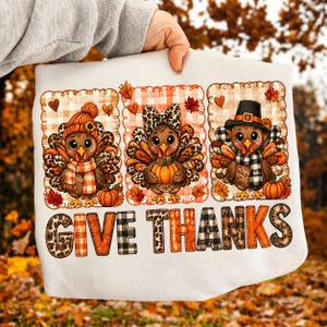 Op de afbeelding: Een wit sweatshirt met drie cartoonkalkoenen in individuele geruite kaders. De kalkoenen dragen hoeden, sjaals en strikken met luipaard- en ruitpatronen. Onder de kalkoenen staat de tekst GIVE THANKS in grote, bedrukte blokletters.