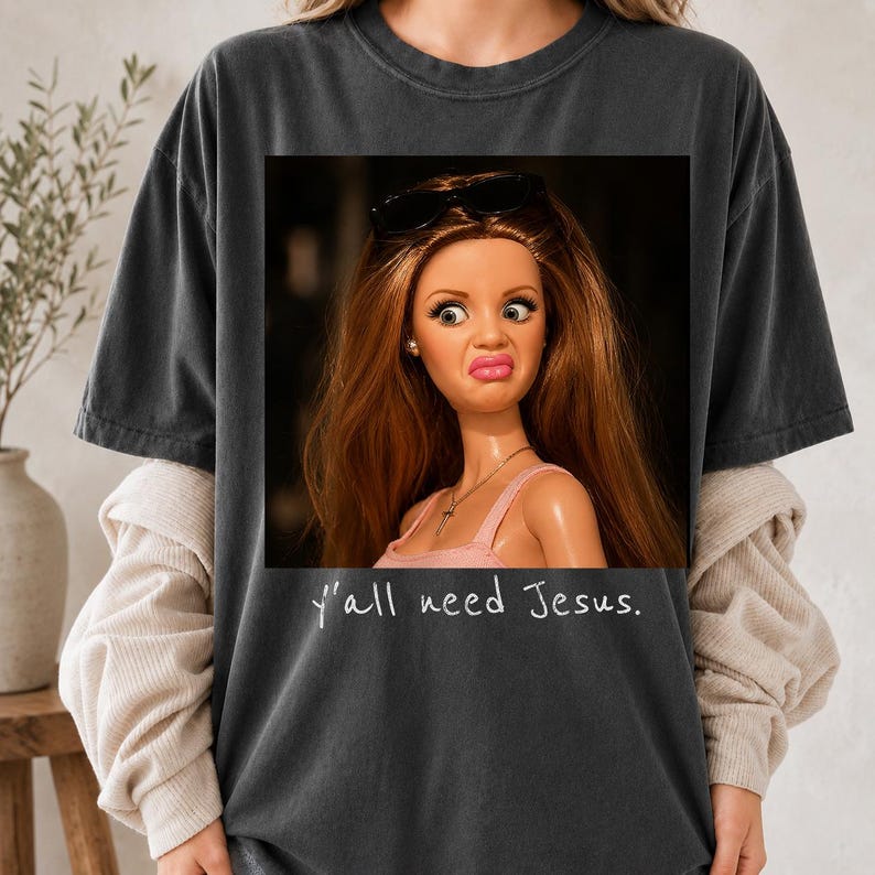 May include: A charcoal gray t-shirt featuring a graphic print of a doll with long brown hair, wearing black sunglasses on her head and a pink top. The doll has a skeptical facial expression. Below the image, the text reads, y'all need Jesus.