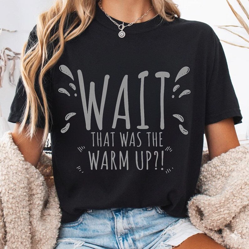 May include: Black t-shirt with the phrase "WAIT THAT WAS THE WARM UP?!" in gray lettering. The shirt has a crew neck and short sleeves. The text is surrounded by splash-like graphics.