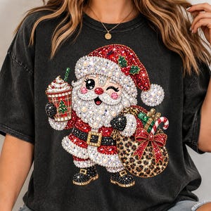 Pode incluir: Uma t-shirt cinzento-carvão com um gráfico do Pai Natal decorado com strass e pérolas brilhantes. O Pai Natal está a piscar o olho, segura um copo xadrez com uma palhinha verde e carrega um saco de presentes com padrão leopardo.