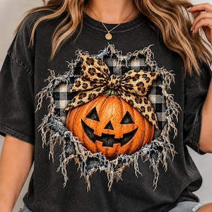 Peut inclure: Un t-shirt gris anthracite avec un motif de citrouille orange sculptée ornée d'un nœud à imprimé léopard. Le design est placé sur un fond à carreaux noir et blanc, encadré par une bordure à l'effet déchiré et effiloché.