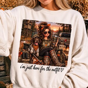 Pode incluir: Uma camisola de gola redonda cor de creme com uma impressão gráfica de uma pessoa com um capuz camuflado, óculos de sol e uma fita para a cabeça, sentada ao lado de um cão com um chapéu camuflado e bandana laranja, com o texto i'm just here for the outfit.