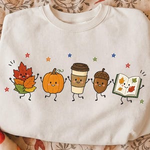 Pode incluir: Uma camisola de gola redonda cor de creme com uma impressão gráfica de cinco personagens antropomórficos: um grupo de folhas de outono, uma abóbora laranja, um copo de café, uma bolota e um livro aberto, rodeados por pequenas estrelas coloridas.