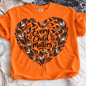 May include: An orange t-shirt featuring a large heart-shaped graphic composed of black, white, and orange feathers, leaves, and small birds. The text "Every Child Matters" is printed in a black script font in the center of the heart design.