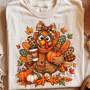 Pode incluir: Uma t-shirt cor de creme com um gráfico de um peru usando um laço xadrez, óculos com estampa de leopardo e um cachecol de tricô. O peru segura um copo de café e uma abóbora, rodeado por folhas de outono, bagas e abóboras adicionais.