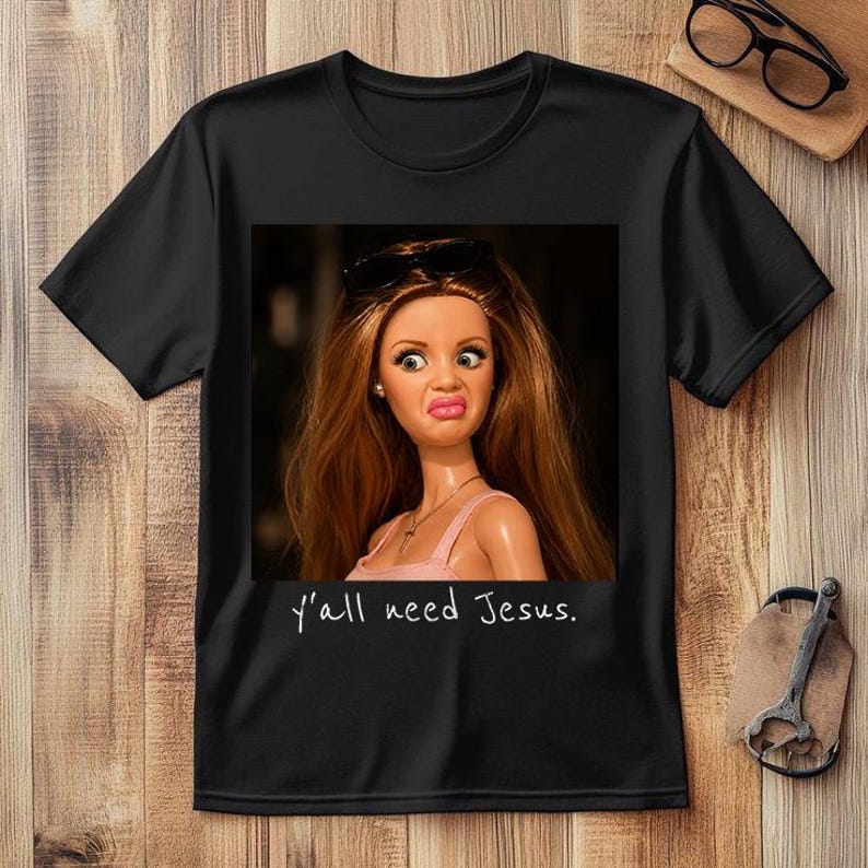 May include: A black short-sleeved t-shirt featuring a graphic print of a doll with long brown hair, wearing sunglasses on her head and a pink top. The doll has a skeptical facial expression. Below the graphic, the text reads, y'all need Jesus.