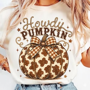 Pode incluir: Uma t-shirt cor de creme com um gráfico de uma abóbora com padrão de manchas de vaca em castanho e branco. A abóbora é rematada com um laço de xadrez vichy castanho e branco. Acima da abóbora, o texto Howdy PUMPKIN está escrito em letras castanhas estilo corda, rodeado por pequenas estrelas castanhas.