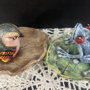 May include: Two decorative ceramic figurines: a bird and a frog. The bird has a gray head, pink face, and orange and yellow wings, perched on a leaf-shaped dish. The frog is blue with red eyes, resting on a green leaf-shaped dish.